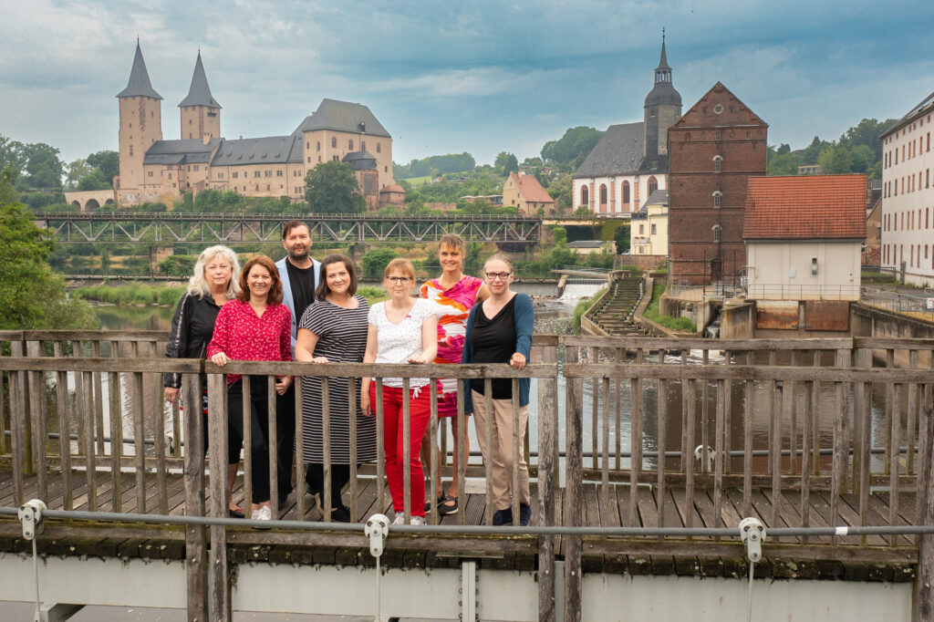 Teamfoto des Regionalmanagements. 7 Personen stehen auf einer Brücke. Im Hintergrund ist das Schloss Rochlitz zu sehen.