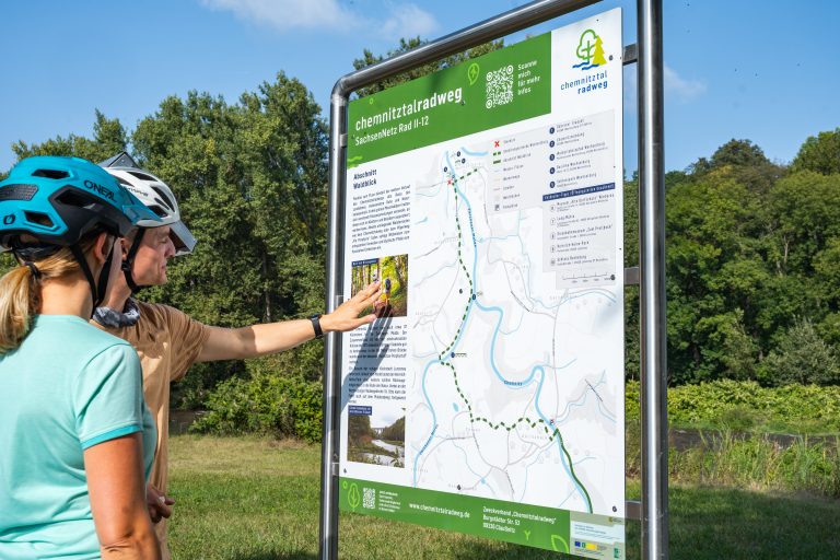 Das Bild zeigt zwei Radfahrer. Sie stehen vor einer Informationstafel und schauen sich den Streckenverlauf des Chemnitztalradweges an.