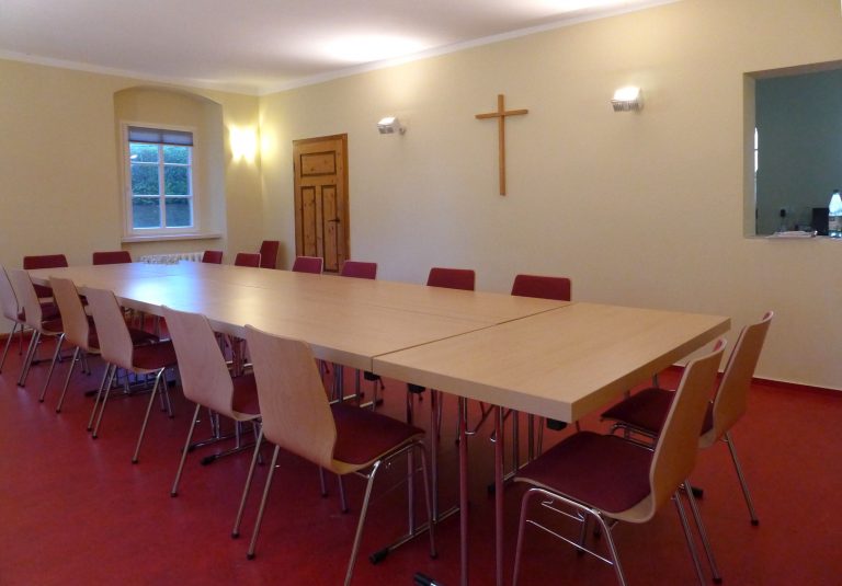 Das Bild zeigt den Gemeinderaum im Pfarrhaus Topfseifersdorf. Im Raum steht ein großer Tisch mit Stühlen drumherum, an der Wand hängt ein Kirchenkreuz.
